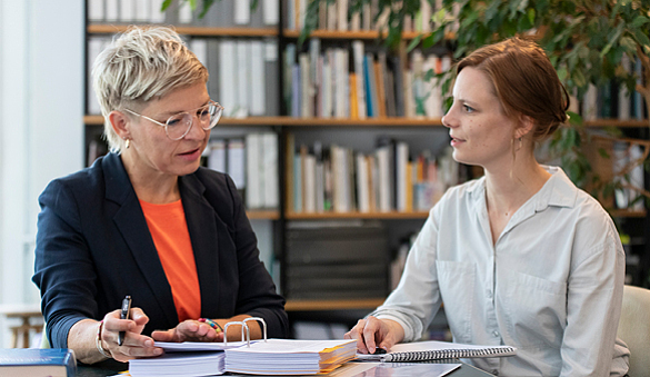 Foto einer Beratungssituation mit zwei Frauen, die an einem Tisch vor einer Bücherwand sitzen