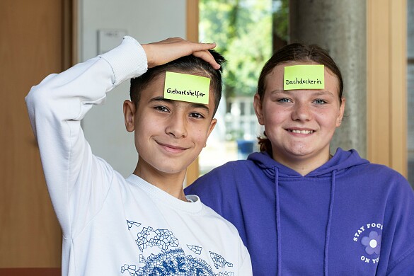 Berufe raten: Abbildung Schüler links mit Zettel auf der Stirn, Geburtshelfer. Rechts daneben, Schülerin mit Zettel auf der Stirn: Dachdeckerin