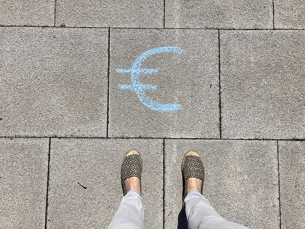 Zwei Frauenbeine die vor einem Eurozeichen stehen, dass mit Kreide auf den Gehweg gemalt ist.